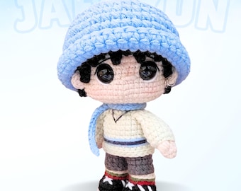 May include: A crocheted amigurumi doll wearing a blue hat, a white sweater, and blue pants. The doll has black hair and large eyes.