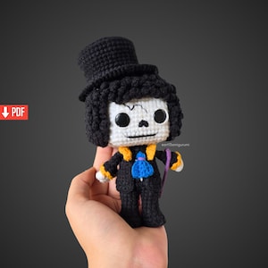 Puede incluir: Una muñeca amigurumi de ganchillo con cabeza de calavera blanca y ojos negros, que lleva un sombrero de copa negro, una chaqueta negra con solapas amarillas y una corbata azul. La figura sostiene un bastón morado e incluye un icono PDF rojo.