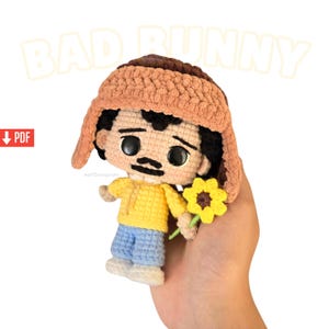 May include: Crocheted doll of a character wearing a brown hat, yellow shirt, and blue pants, holding a yellow flower. The doll has a black mustache and black hair. The word "BAD BUNNY" is in the background.