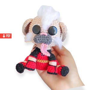 May include: A handmade crochet dog doll with large black eyes, a gray muzzle, and a pink tongue sticking out. The dog wears a red and black outfit with gold accents. The doll is held in a hand.