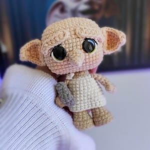 May include: A crocheted amigurumi doll of a house elf with large eyes, wearing a white dress and holding a grey object. The doll is light brown and beige.
