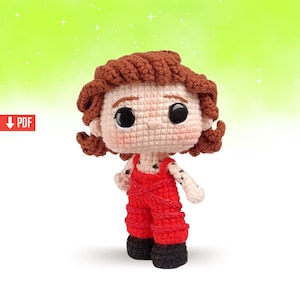 May include: Crochet amigurumi doll with brown hair, wearing red dungarees and black shoes. The doll has large black eyes and a small, pink nose.