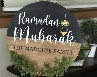 Puede incluir: Un letrero redondo de madera con un acabado de madera de grano negro y marrón. El letrero dice "Ramadan Mubarak" en letras blancas en cursiva con un gráfico de linterna dorada. La parte inferior del letrero dice "The Madouse Family" en letras negras.