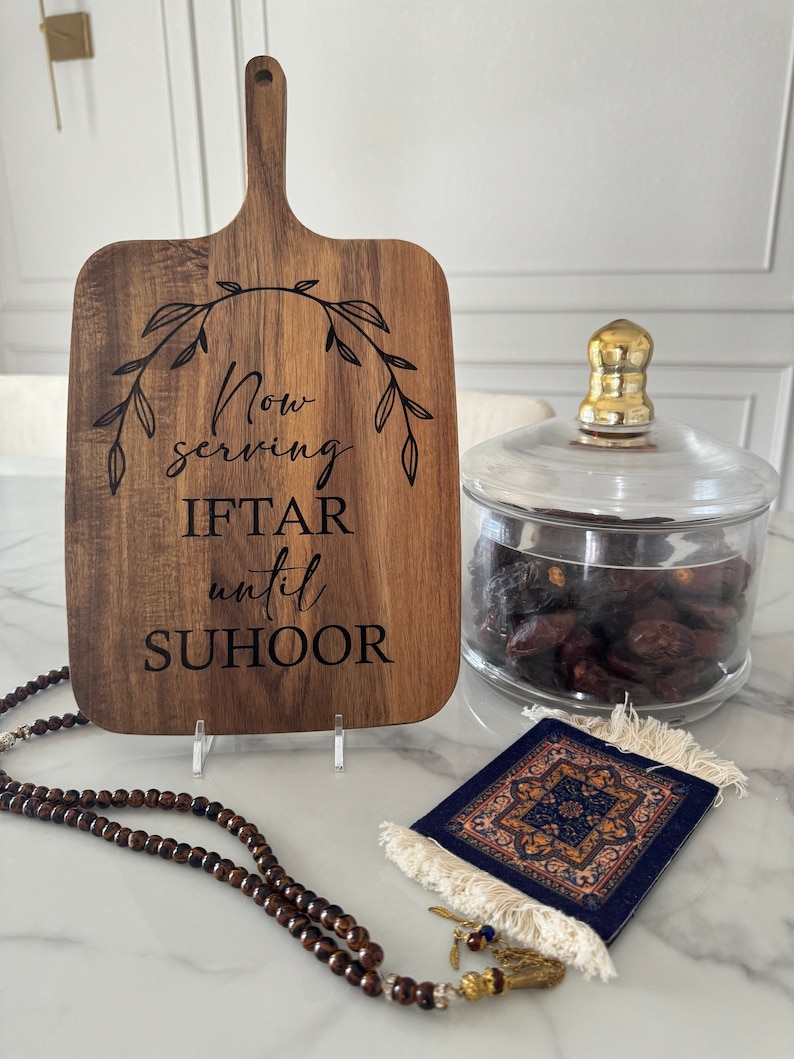A wooden cutting board with text next to a bowl of dates, a small prayer rug, and a beaded prayer necklace on a counter