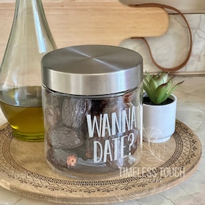 May include: A glass jar with a silver lid filled with dates. The jar has a white sticker that says "Wanna Date?" on it. The jar is sitting on a wooden tray with a decorative pattern.