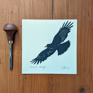 May include: A black and white print of a bird in flight, with the words "Cornish chough" and a signature. A wood carving tool with a wooden handle is to the left of the print. The print is on a cream-coloured paper.