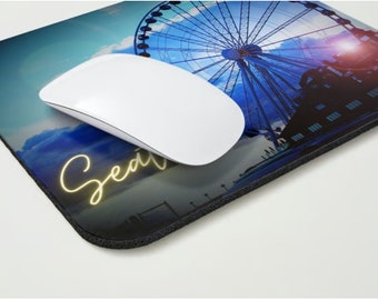 May include: A white computer mouse rests on a mousepad featuring a blue sky, clouds, and a Ferris wheel. The word "Sea" is written in a glowing yellow font on the mousepad.