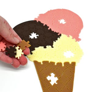 Peut inclure: Une main tient des pièces de puzzle d'un cône de glace au chocolat, à la vanille et à la fraise. Les pièces manquent au puzzle du cône de glace plus grand.