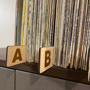 Puede incluir: Dos separadores de madera etiquetados con "A" y "B" se utilizan para organizar una colecci&oacute;n de discos de vinilo. Los discos est&aacute;n apilados verticalmente y tienen varios t&iacute;tulos y artistas impresos en sus lomos.