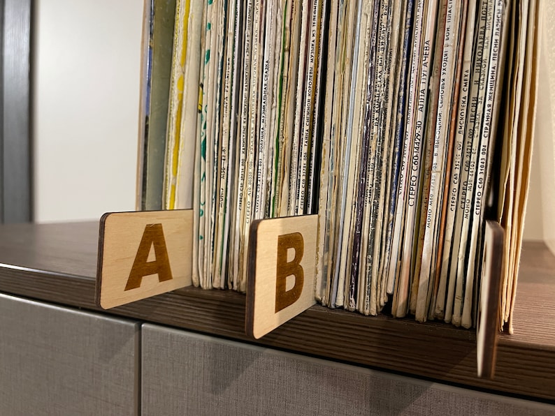 Puede incluir: Dos separadores de madera etiquetados con "A" y "B" se utilizan para organizar una colecci&oacute;n de discos de vinilo. Los discos est&aacute;n apilados verticalmente y tienen varios t&iacute;tulos y artistas impresos en sus lomos.