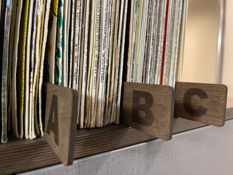 Puede incluir: Primer plano de un estante de madera con una colecci&oacute;n de discos de vinilo. Los discos est&aacute;n organizados por letra, con separadores de madera etiquetados con "A", "B" y "C".