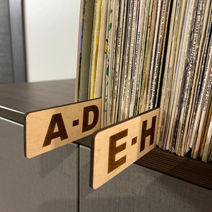 Puede incluir: Dos etiquetas de madera con el texto "A-D" y "E-H" est&aacute;n unidas a un estante con una colecci&oacute;n de discos de vinilo.