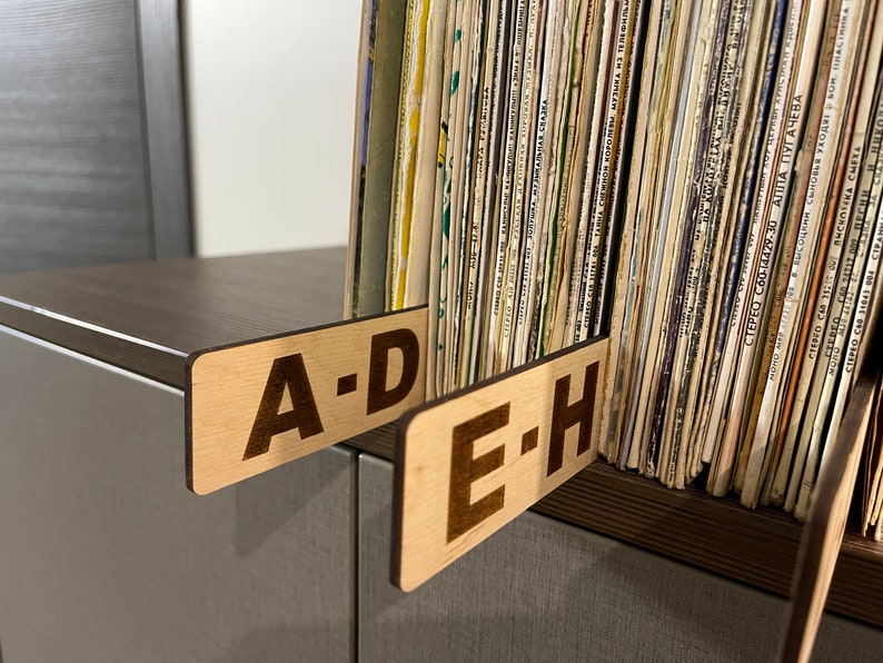 Puede incluir: Dos etiquetas de madera con el texto "A-D" y "E-H" est&aacute;n unidas a un estante con una colecci&oacute;n de discos de vinilo.