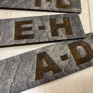 Puede incluir: Tres letreros de madera con las letras "A-D", "E-H" y "I-L" grabadas en la superficie. Los letreros son de color marr&oacute;n oscuro con un patr&oacute;n de veta de madera.