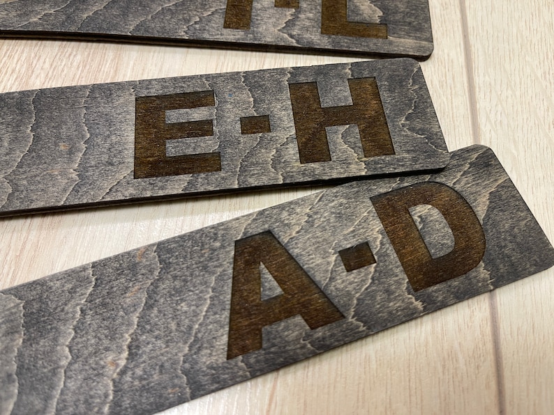 Puede incluir: Tres letreros de madera con las letras "A-D", "E-H" y "I-L" grabadas en la superficie. Los letreros son de color marr&oacute;n oscuro con un patr&oacute;n de veta de madera.