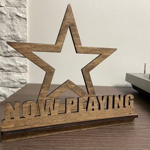 May include: A wooden star-shaped sign with the words "NOW PLAYING" cut out. The sign is on a wooden base and has a dark brown stain.