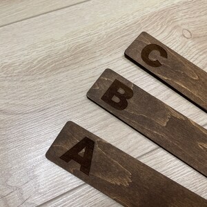 Puede incluir: Tres marcap&aacute;ginas de madera con las letras A, B y C grabadas en ellos. Los marcap&aacute;ginas son de color marr&oacute;n oscuro y tienen un patr&oacute;n de veta de madera.