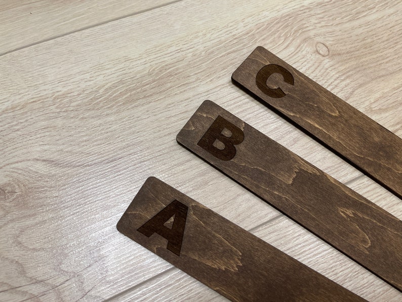 Puede incluir: Tres marcap&aacute;ginas de madera con las letras A, B y C grabadas en ellos. Los marcap&aacute;ginas son de color marr&oacute;n oscuro y tienen un patr&oacute;n de veta de madera.