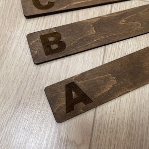 Puede incluir: Tres marcap&aacute;ginas de madera con las letras A, B y C grabadas en ellos. Los marcap&aacute;ginas son de color marr&oacute;n y tienen una veta de madera natural.