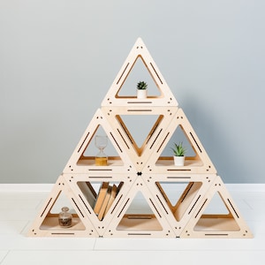 May include: A light-colored wooden triangular shelf unit, assembled in a pyramid shape. The shelves contain small potted plants, books, a decorative bottle, and an hourglass. The unit is on a white floor against a gray wall.