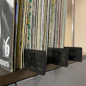Puede incluir: Primer plano de una estanter&iacute;a de discos de vinilo, organizados por letra. La estanter&iacute;a es de madera oscura y tiene tres separadores etiquetados con "A", "B" y "C".