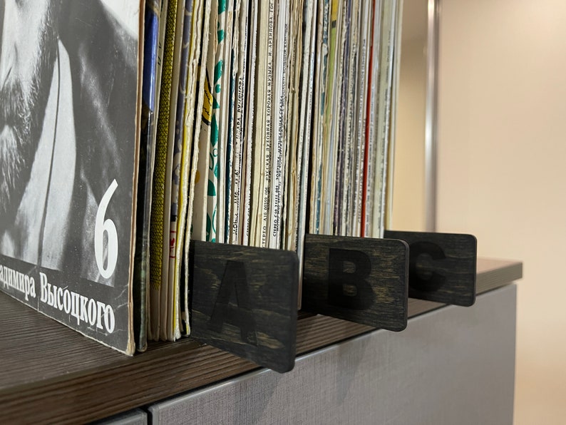 Puede incluir: Primer plano de una estanter&iacute;a de discos de vinilo, organizados por letra. La estanter&iacute;a es de madera oscura y tiene tres separadores etiquetados con "A", "B" y "C".