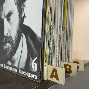 Puede incluir: Primer plano de una colecci&oacute;n de discos de vinilo con separadores de madera etiquetados con A, B y C. El disco de la izquierda tiene una foto en blanco y negro de un hombre con barba y el texto "На концертах Владимира Высоцкого 6".