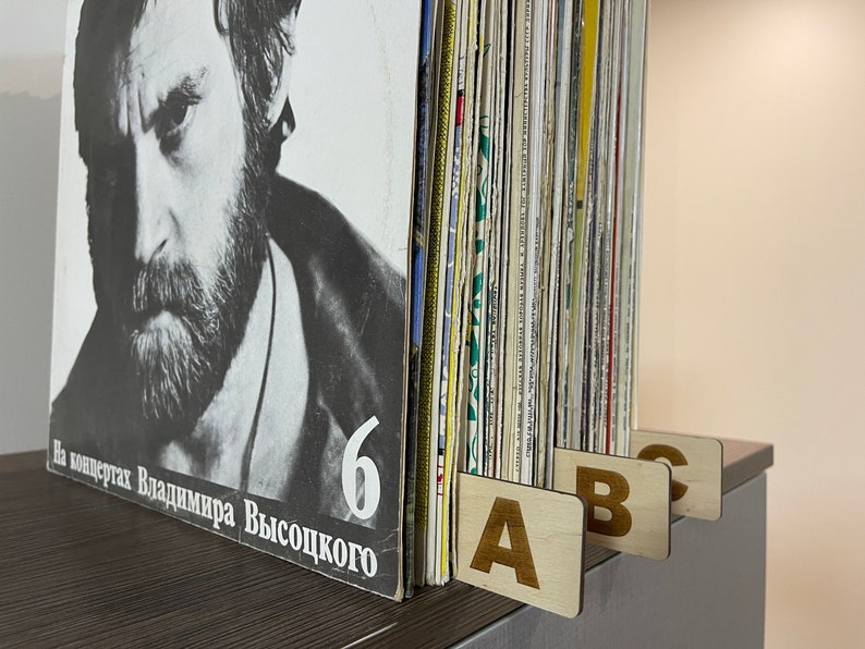 Puede incluir: Primer plano de una colecci&oacute;n de discos de vinilo con separadores de madera etiquetados con A, B y C. El disco de la izquierda tiene una foto en blanco y negro de un hombre con barba y el texto "На концертах Владимира Высоцкого 6".