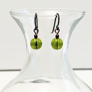 May include: A pair of dangle earrings with olive green glass beads and dark copper-colored hooks. The round beads have a faceted design, and the earrings are displayed against a blurred glass background.