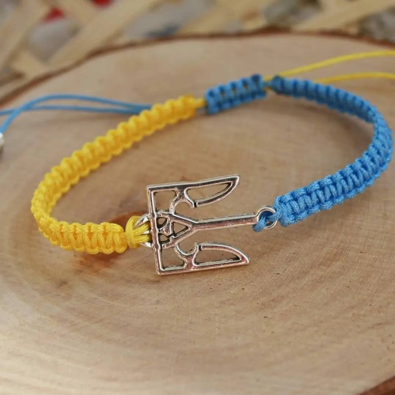 May include: A handmade bracelet featuring a silver trident charm. The bracelet is made of yellow and blue braided cord, with a sliding knot closure. The colors are reminiscent of a national flag. The bracelet is displayed on a wooden surface.