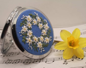 Puede incluir: Un espejo compacto plateado con una cubierta de tela azul bordada con una corona de margaritas blancas. El espejo está sobre una hoja de música con una flor de narciso amarilla.