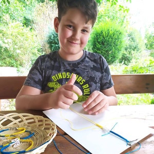 May include: A young person smiles while working on a macrame project. A basket of finished blue and yellow bracelets sits on a wooden table. The person is wearing a gray camouflage t-shirt with the text "DINOSAURS RULE".