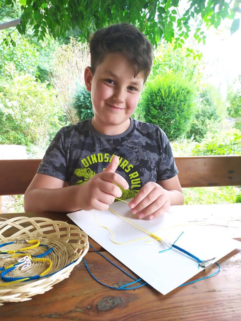 May include: A young person smiles while working on a macrame project. A basket of finished blue and yellow bracelets sits on a wooden table. The person is wearing a gray camouflage t-shirt with the text "DINOSAURS RULE".