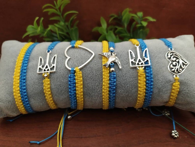 May include: A collection of macrame bracelets in blue and yellow, featuring various silver charms. The charms include a heart, a bird, and a trident. The bracelets are displayed on a gray pillow, with green foliage in the background.
