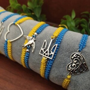 May include: Macrame bracelets in blue and yellow, featuring silver charms. Charms include a trident, a heart, and a bird. The bracelets are displayed on a gray surface, with green foliage in the background.