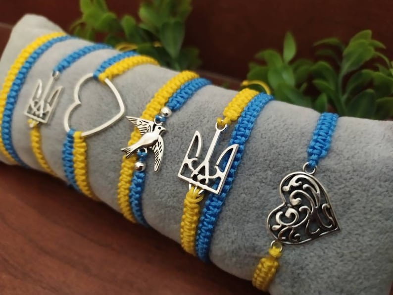 May include: Macrame bracelets in blue and yellow, featuring silver charms. Charms include a trident, a heart, and a bird. The bracelets are displayed on a gray surface, with green foliage in the background.