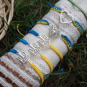 May include: A collection of handmade bracelets in blue and yellow, with silver charms. The bracelets are displayed on a white and brown knitted surface, set against a backdrop of green grass. Some bracelets feature a heart or trident charm.