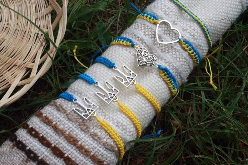 May include: A collection of handmade bracelets in blue and yellow, with silver charms. The bracelets are displayed on a white and brown knitted surface, set against a backdrop of green grass. Some bracelets feature a heart or trident charm.