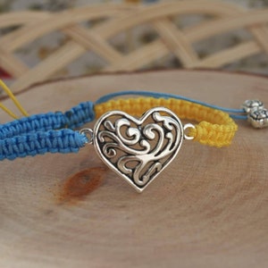 May include: Macrame bracelets in blue and yellow, featuring a silver heart-shaped charm with an intricate design. The bracelets are displayed on a wooden surface, with decorative silver beads.