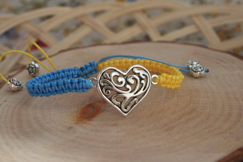 May include: Macrame bracelets in blue and yellow, featuring a silver heart-shaped charm with an intricate design. The bracelets are displayed on a wooden surface, with decorative silver beads.