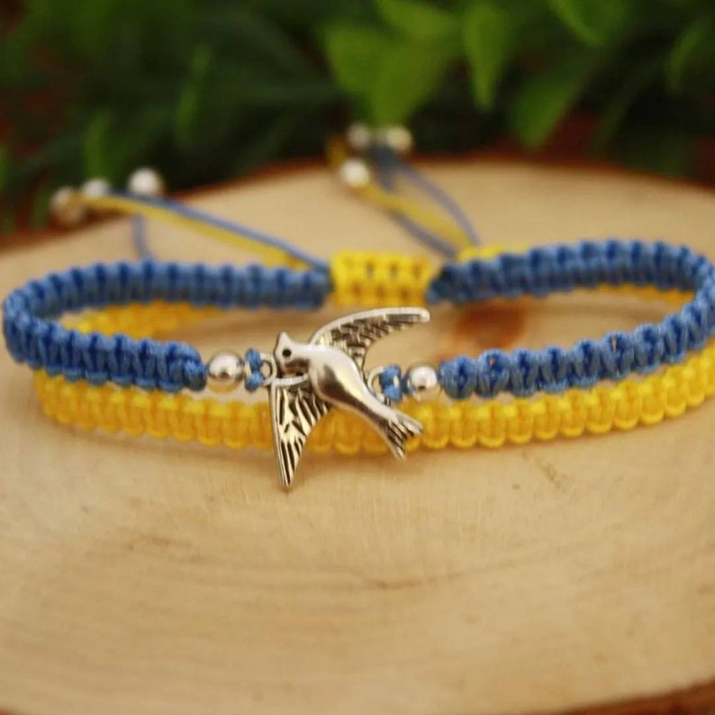 May include: Macrame bracelet in blue and yellow, featuring a silver bird charm and silver beads. The bracelet is adjustable and sits on a wooden surface. The colors are reminiscent of a flag.