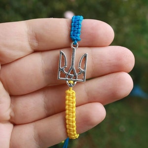 May include: A silver pendant in the shape of the Ukrainian Tryzub, suspended between blue and yellow braided cords. The bracelet is held in a hand against a blurred green background.