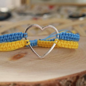 May include: A macrame bracelet featuring a silver heart charm. The bracelet is made with blue and yellow macrame, with the colors arranged in stripes. The bracelet is displayed on a wooden surface.