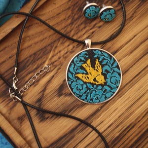 May include: A black cord necklace with a round pendant featuring a yellow bird embroidered on a blue floral pattern. The pendant is attached to a silver chain. The necklace comes with a pair of matching earrings.