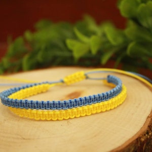 May include: A handmade macrame bracelet in the colors of the Ukrainian flag: yellow and blue. The bracelet is displayed on a wooden slice with green foliage in the background. The bracelet is adjustable.
