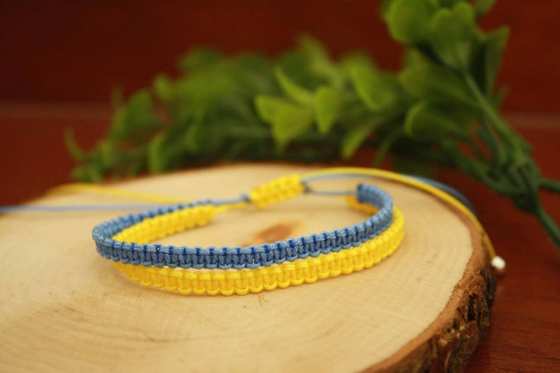 May include: A handmade macrame bracelet in the colors of the Ukrainian flag: yellow and blue. The bracelet is displayed on a wooden slice with green foliage in the background. The bracelet is adjustable.