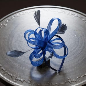 May include: A royal blue headband with a decorative bow and gray feathers. The bow is made of looped fabric, and the feathers are attached to the top. The headband is on a silver surface.