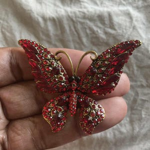 May include: A red butterfly brooch with gold-colored accents. The brooch is covered in small, sparkling red crystals. The butterfly's wings are spread, and the antennae are curved. A decorative accessory.