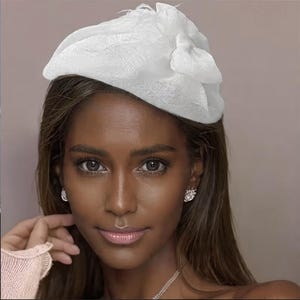May include: A white, decorative hat with a bow and feather accents. The hat is made of a soft, textured fabric. The hat is worn by a person with brown hair and dark eyes. The person is wearing diamond earrings and a necklace.