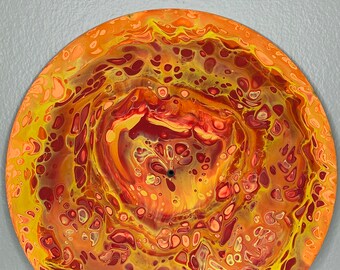 Puede incluir: Obra de arte circular con un diseño abstracto en tonos cálidos. La pieza presenta un patrón arremolinado de amarillos, naranjas y rojos. La obra tiene un agujero central, lo que sugiere que podría ser una esfera de reloj o un elemento decorativo.
